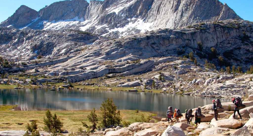 A group of outward bound students hike along rocky terrain near an alpine lake. There is a large mountain in the background. 